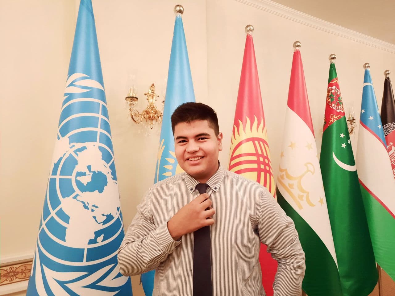 A man stands in front of flags. He is wearing a dress shirt and tie and smiling. 